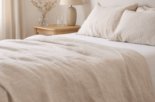 Soft, neutral-toned bedroom with a neatly made linen-covered bed, wooden bedside table, lamp and small vase of dried flowers in natural daylight, creating a calm and restful atmosphere.