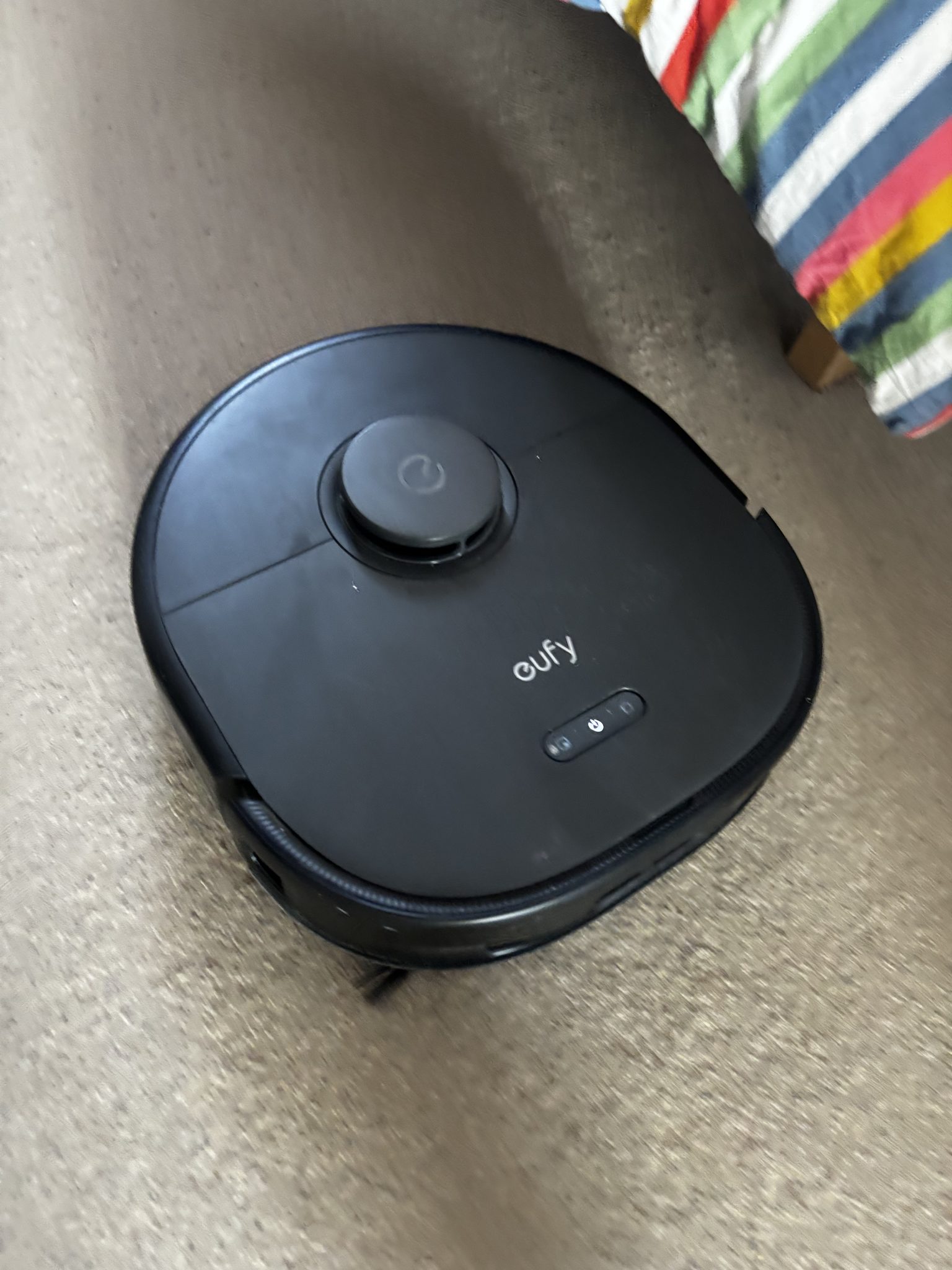 A eufy X10 Pro Omni robot vacuum part-way through a cleaning run on a carpeted living-room floor, beside a dehumidifier and a striped throw