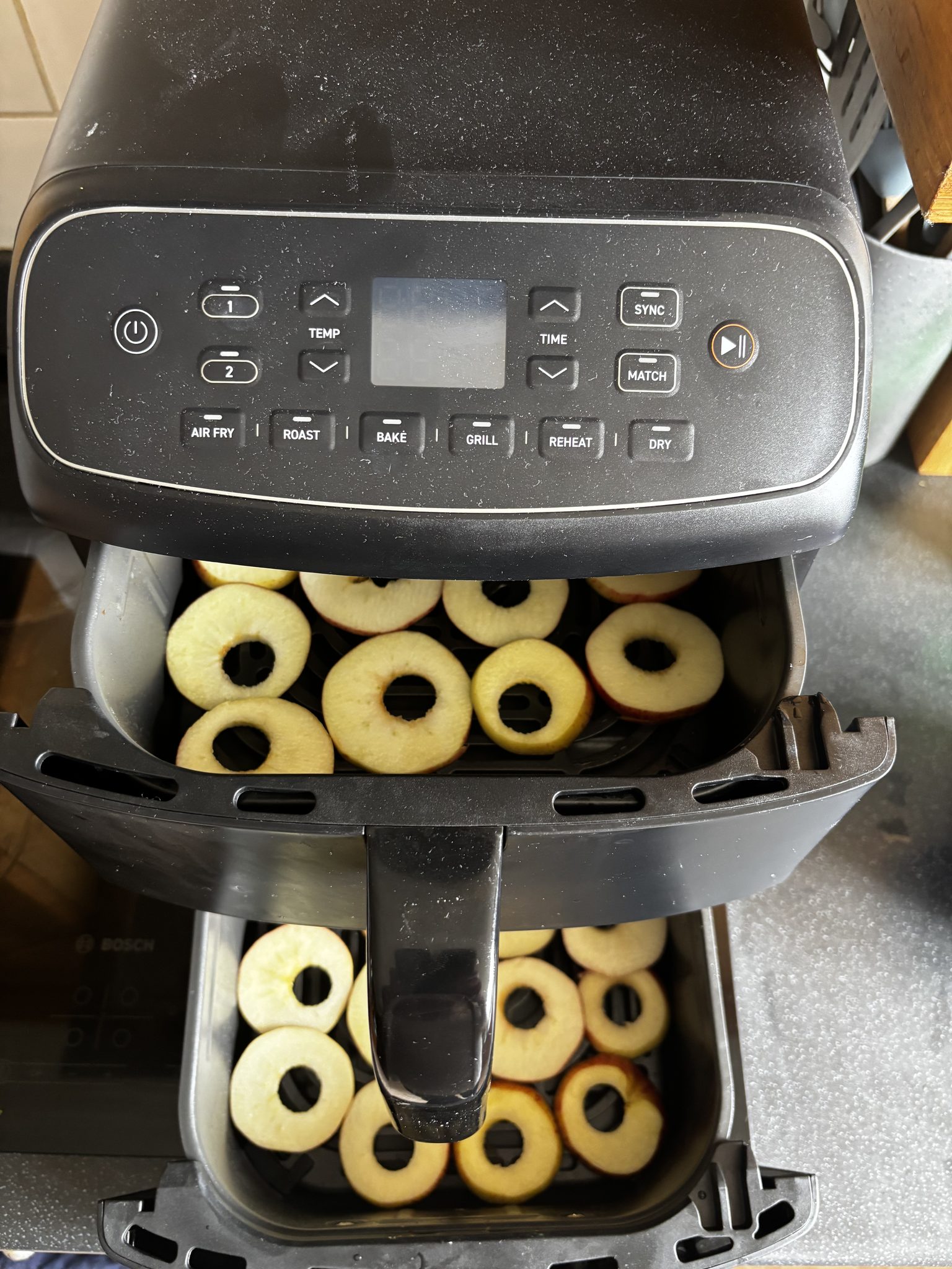 Both drawers of a two-drawer air fryer open, each holding about a dozen apple rings in a single layer; the control panel above shows AIR FRY, ROAST, BAKE, GRILL, REHEAT and DRY.