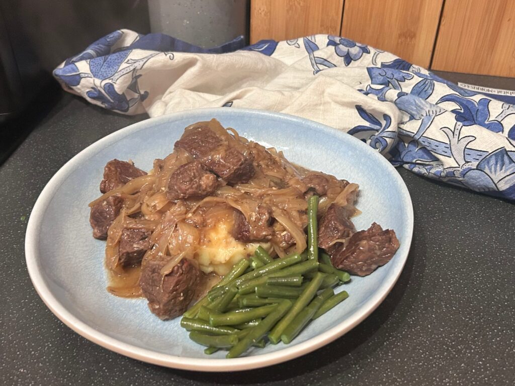 Beef carbonnade with slow-cooked onions spooned over mash, with green beans, on a pale blue plate in front of a blue and white tea towel