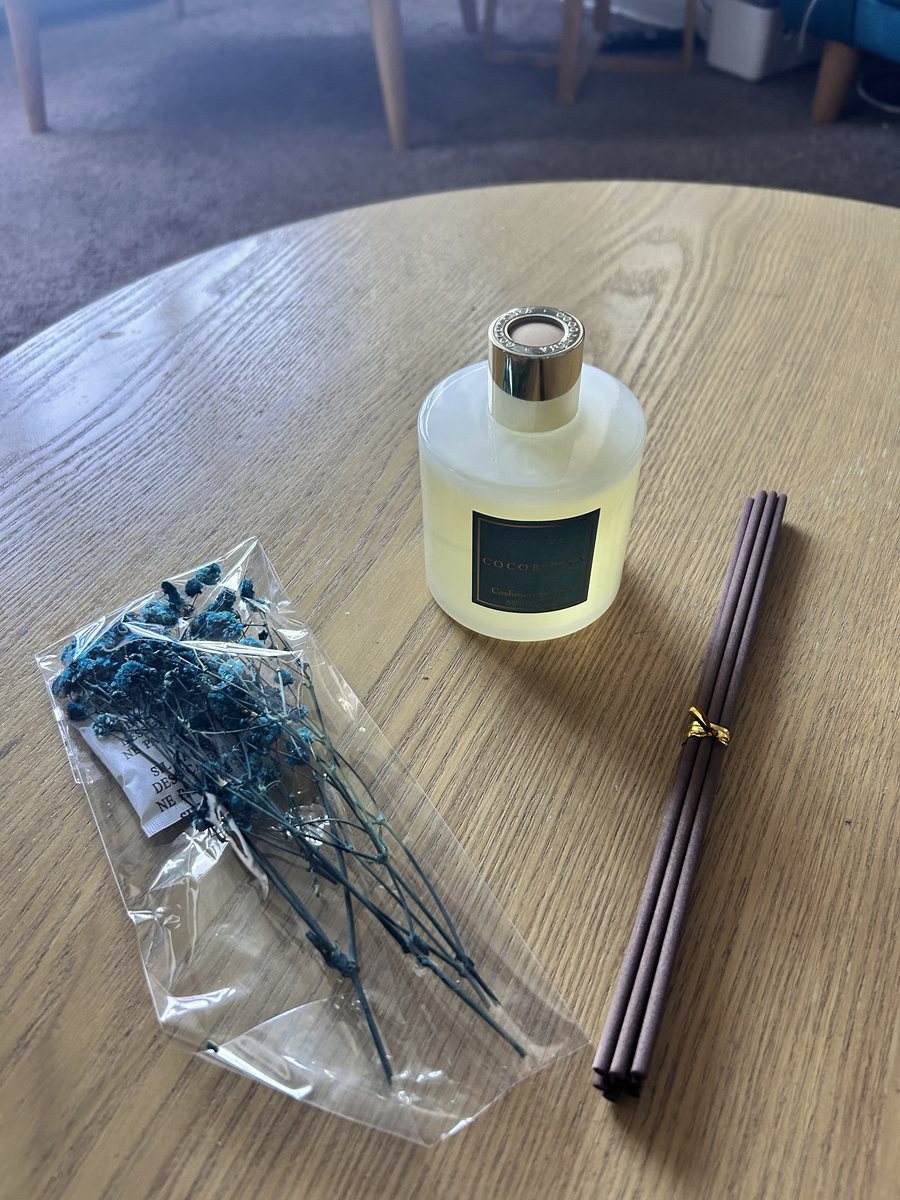 The Cocorrína Cashmere Vanilla reed diffuser unboxed on a wooden table: a 200ml frosted glass bottle, a bundle of brown fibre reeds tied with gold ribbon, and a bag of dried blue flowers