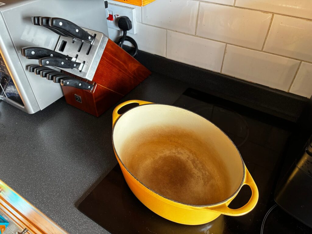 A yellow enamelled cast-iron oval casserole with the lid off, showing the cream enamel interior browned with use, on an induction hob beside a knife block