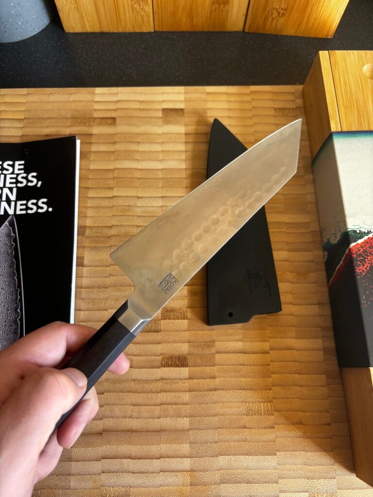 A hand holding the Kotai bunka knife over a bamboo chopping board, showing the hammered finish on the blade