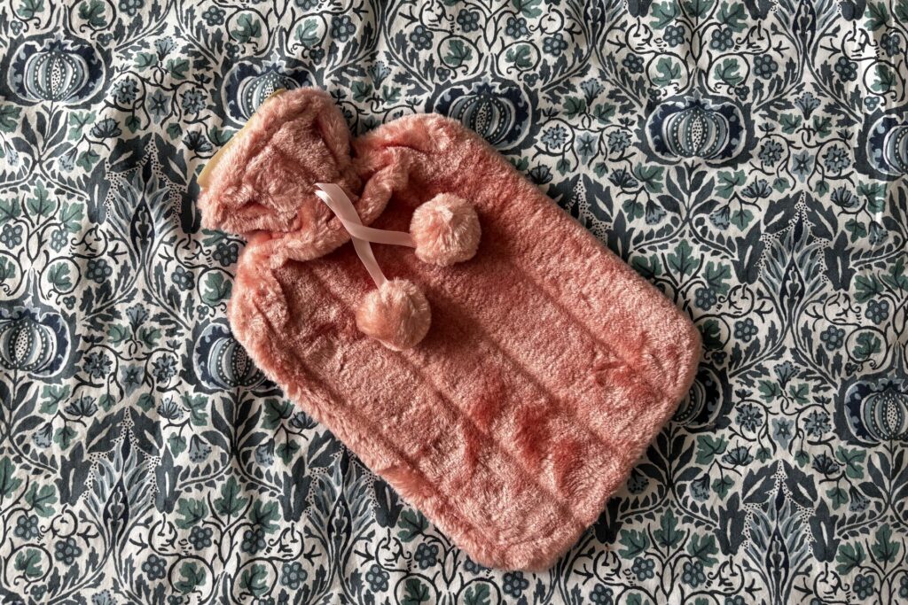 A pink faux fur covered hot water bottle with pom-pom ties on a patterned duvet