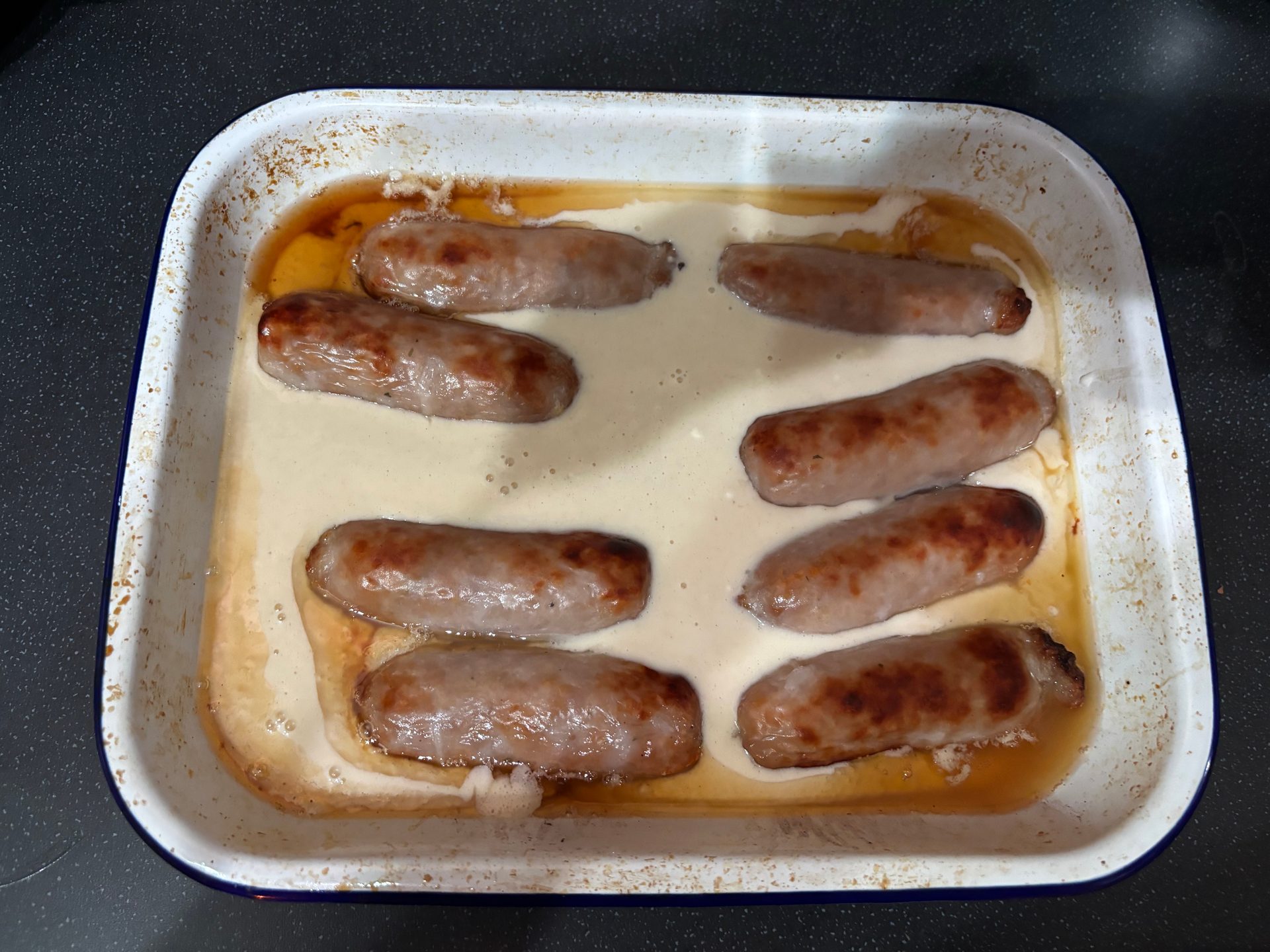 Browned sausages in a white enamel roasting tray with the batter just poured in around them